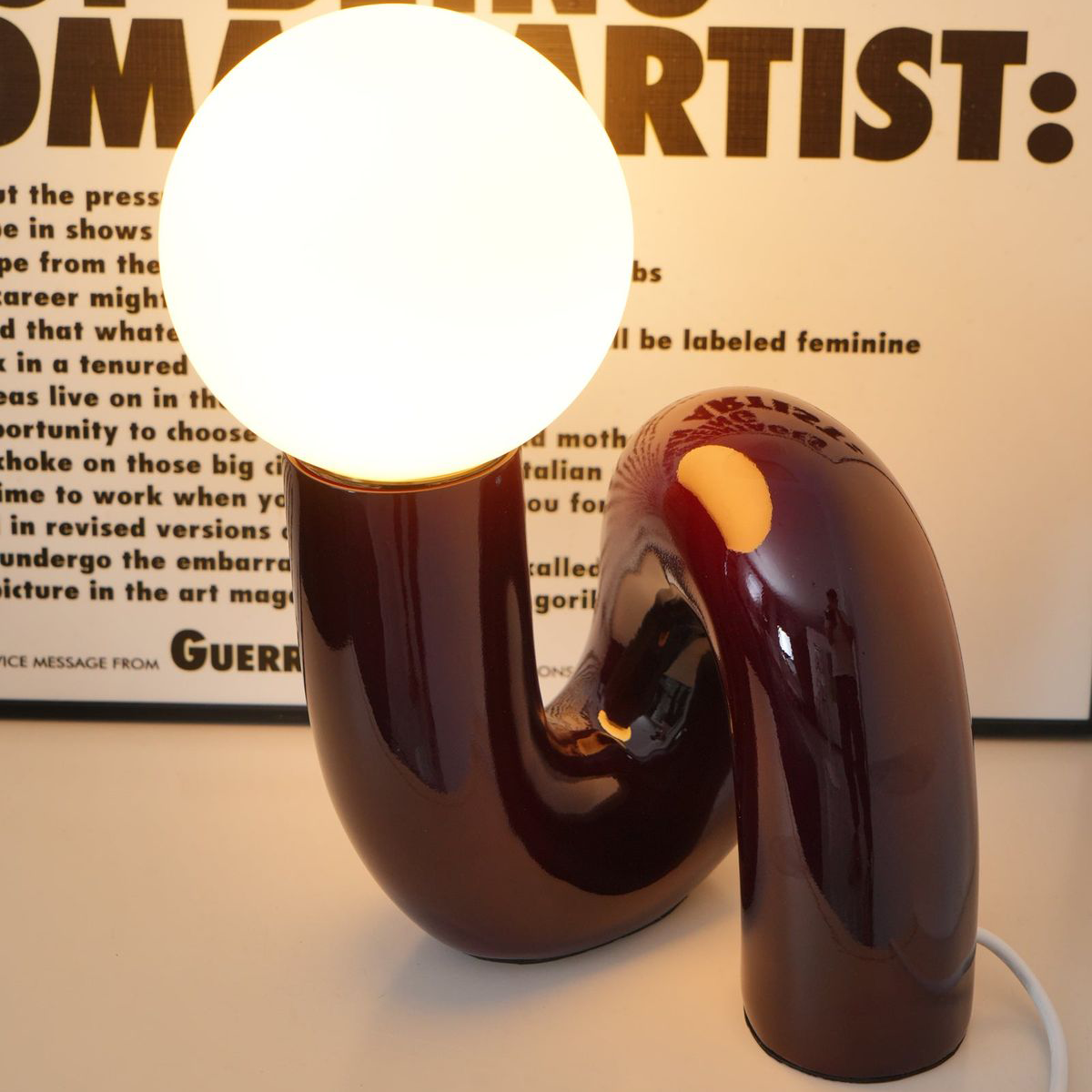 A Burgundy Red Gittana Table Lamp glowing in front of a typographic poster