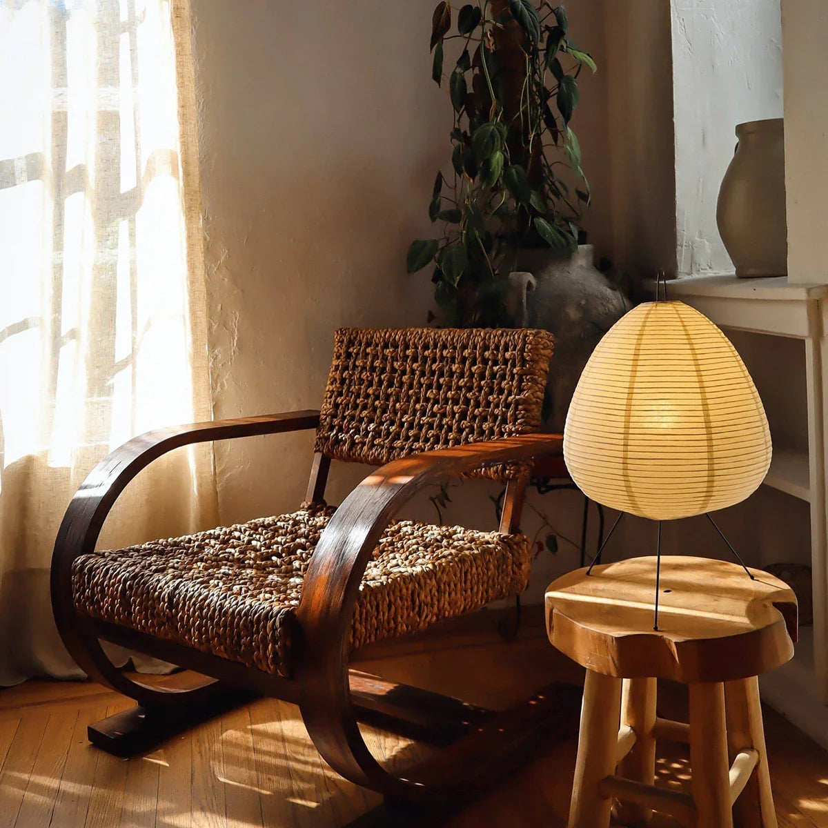 A Hachi Paper Lamp glowing on a wooden stool beside a rattan armchair
