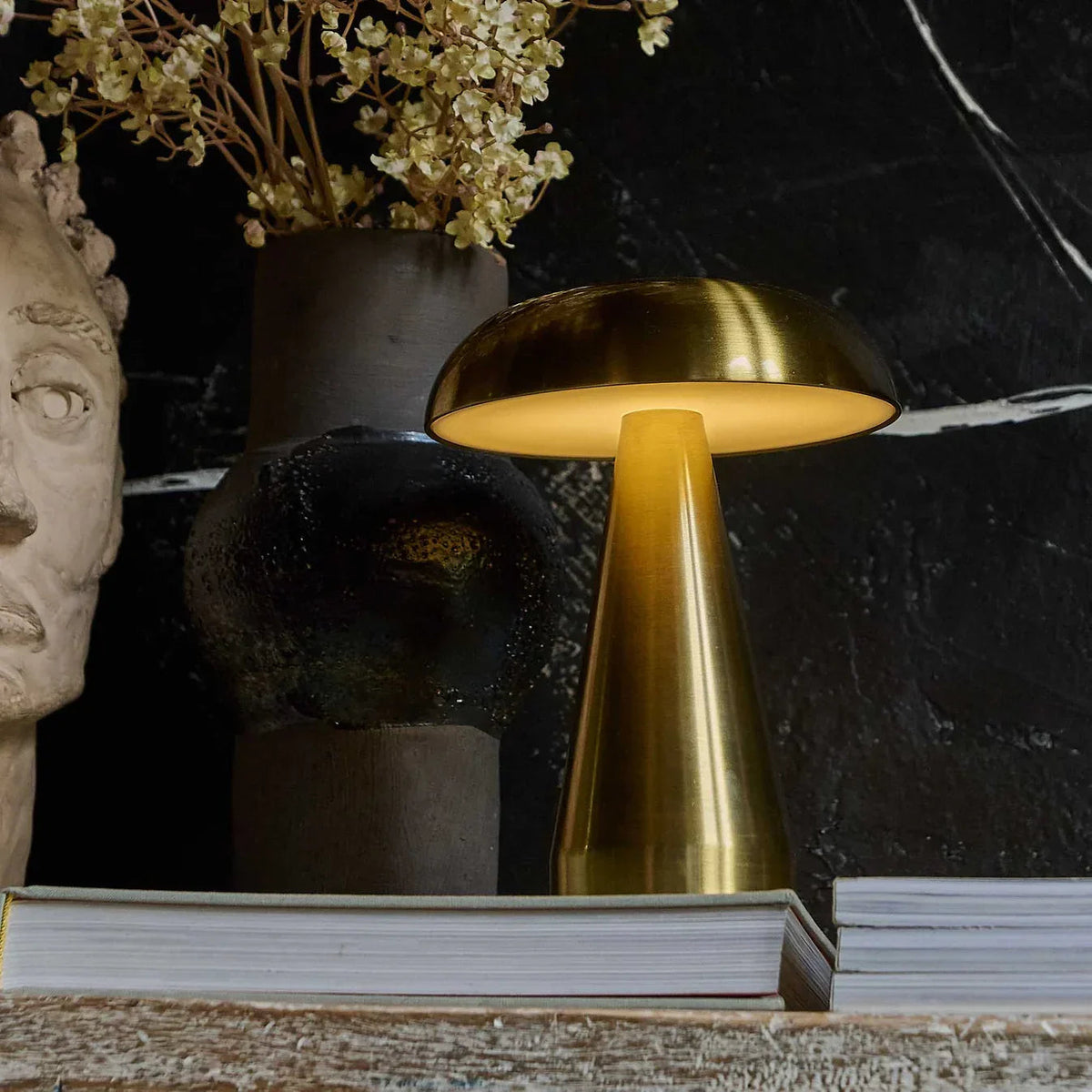 A Brushed Gold Lucido Portable Lamp glowing on a dark marble surface beside a sculpture