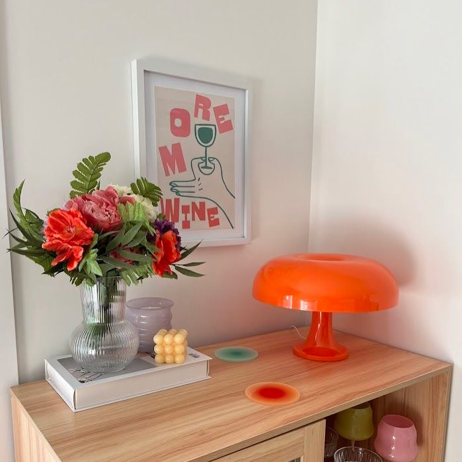 A Tangerine Orange Strada Table Lamp on a wooden sideboard beside a vase of red flowers