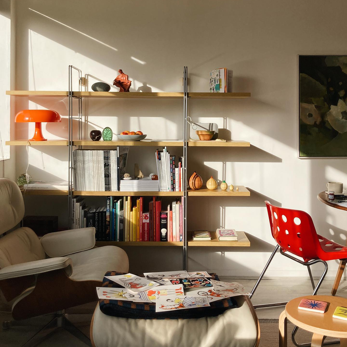 A Tangerine Orange Strada Table Lamp on open shelving in a sunny living room with a red chair