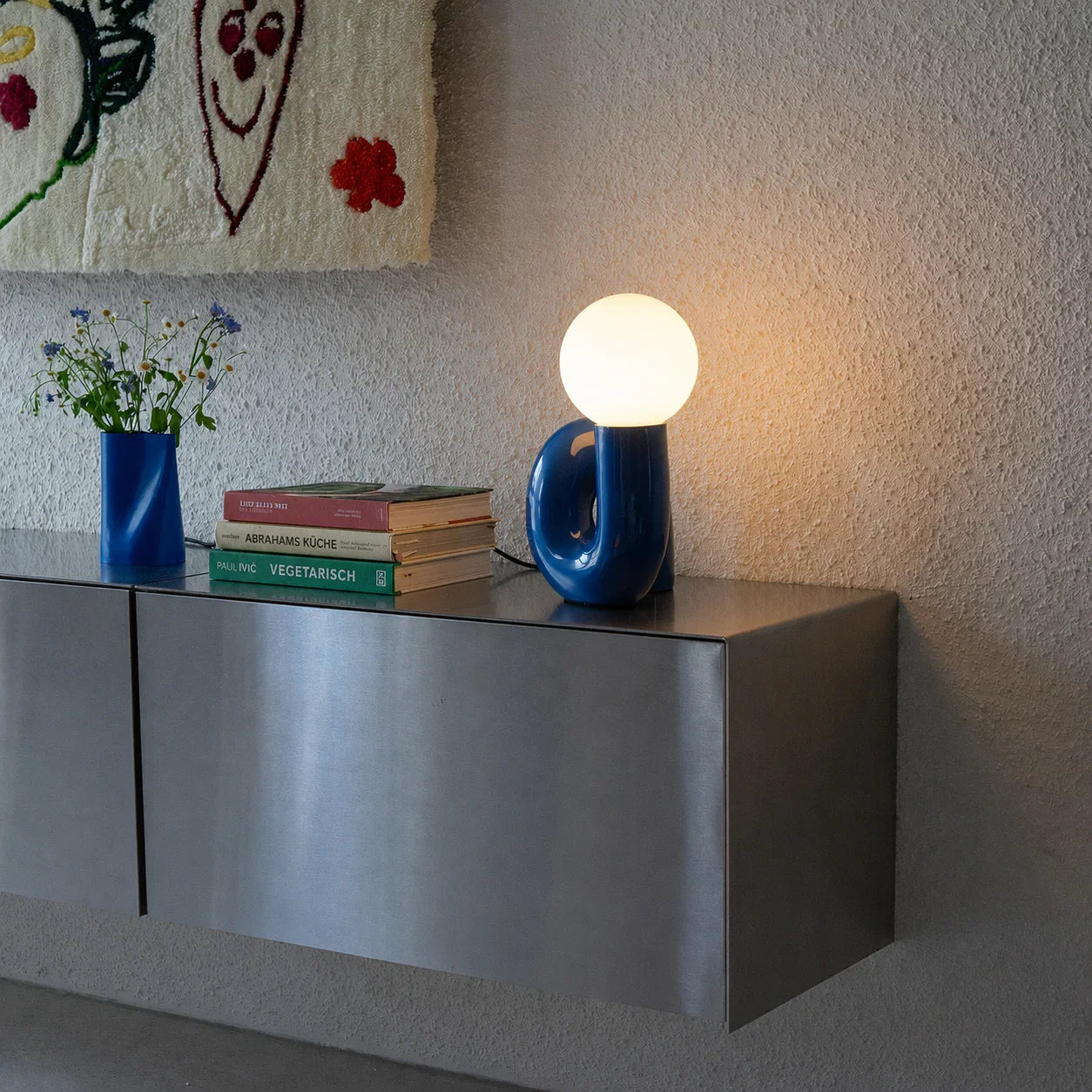 Blue modern table lamp with round bulb on metal shelf, styled with books and vase