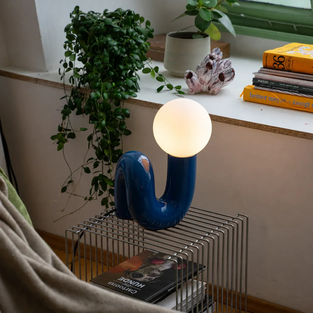 Groovy blue curved lamp with glowing orb, styled on wire shelf with plants and books