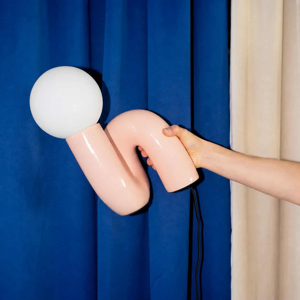 Pink curved table lamp with white globe shade held against blue curtain, funky modern lighting