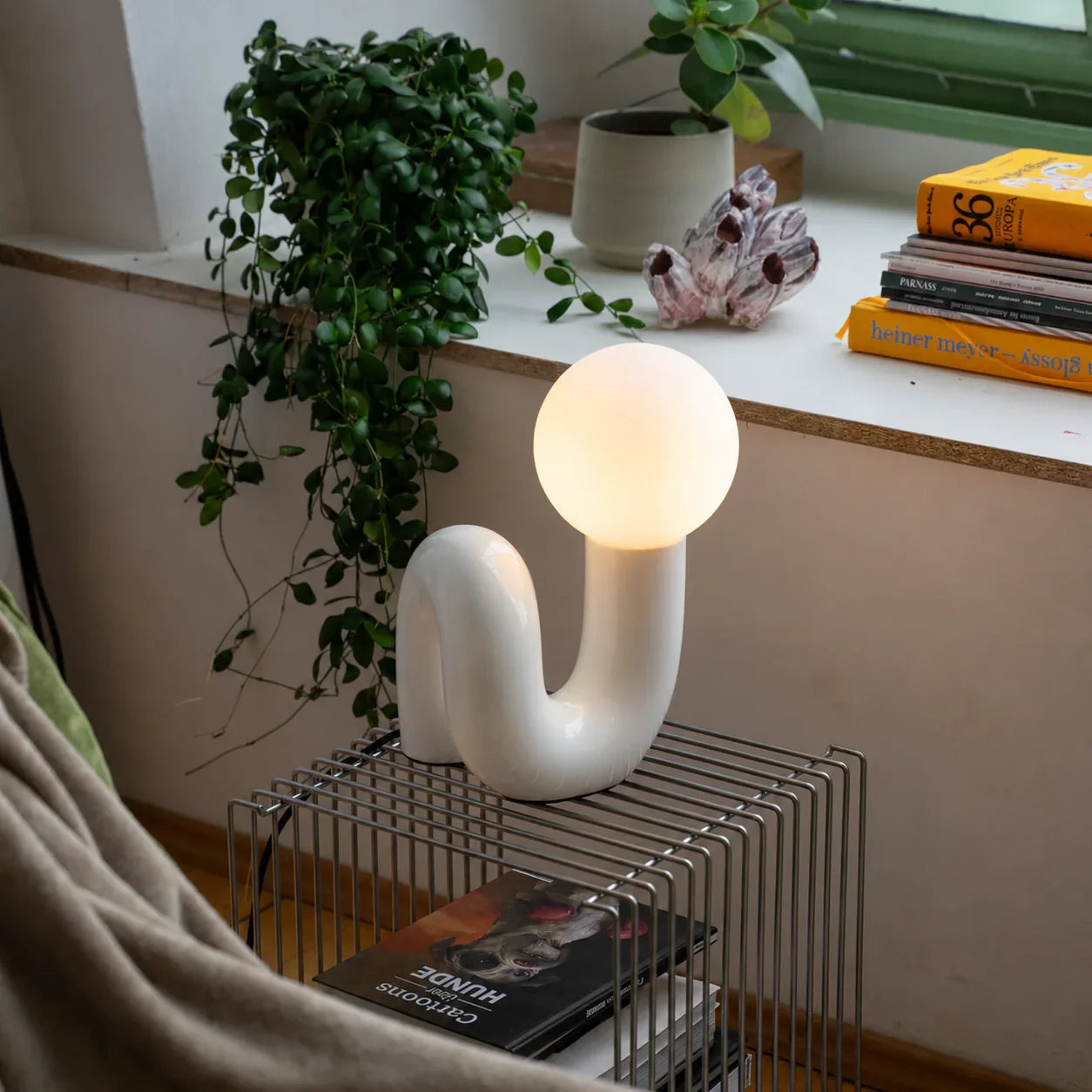 Groovy white curved lamp with glowing orb, styled on wire shelf with plants and books
