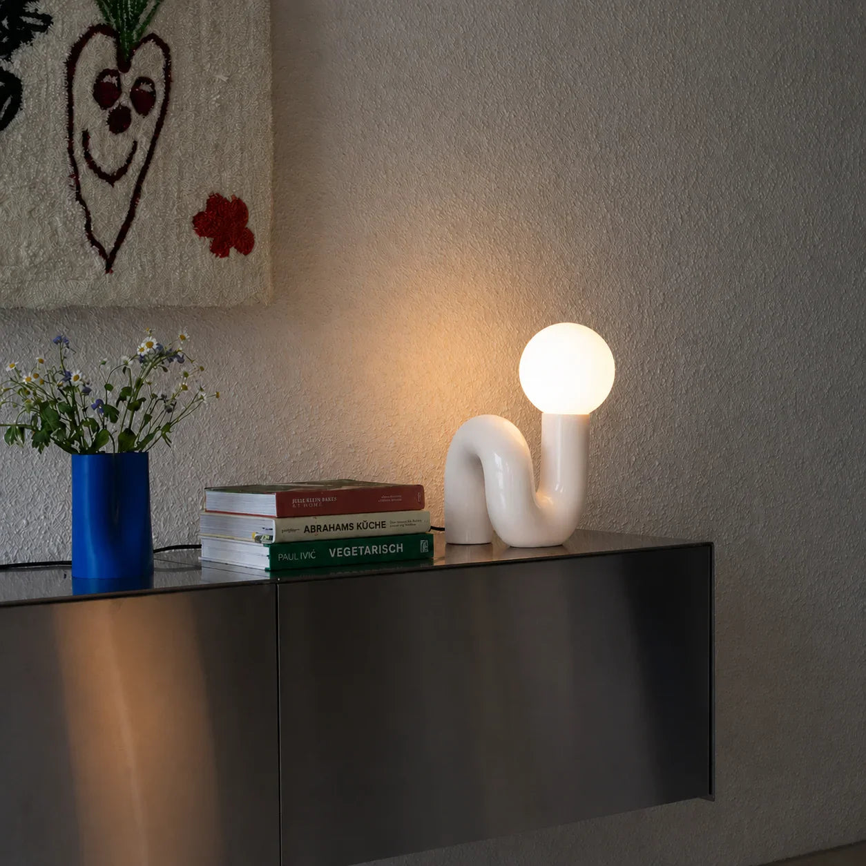 Modern white curved lamp with glowing bulb on sleek console table beside books and flowers