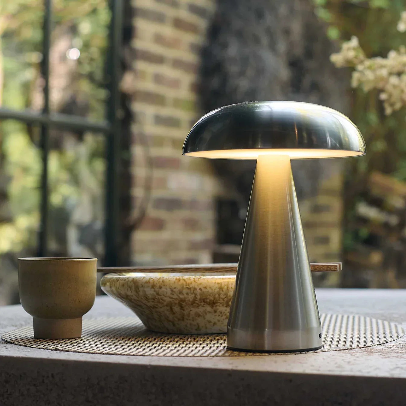 Modern mushroom-style table lamp with warm glow, styled on a textured mat beside ceramic bowl and cup.