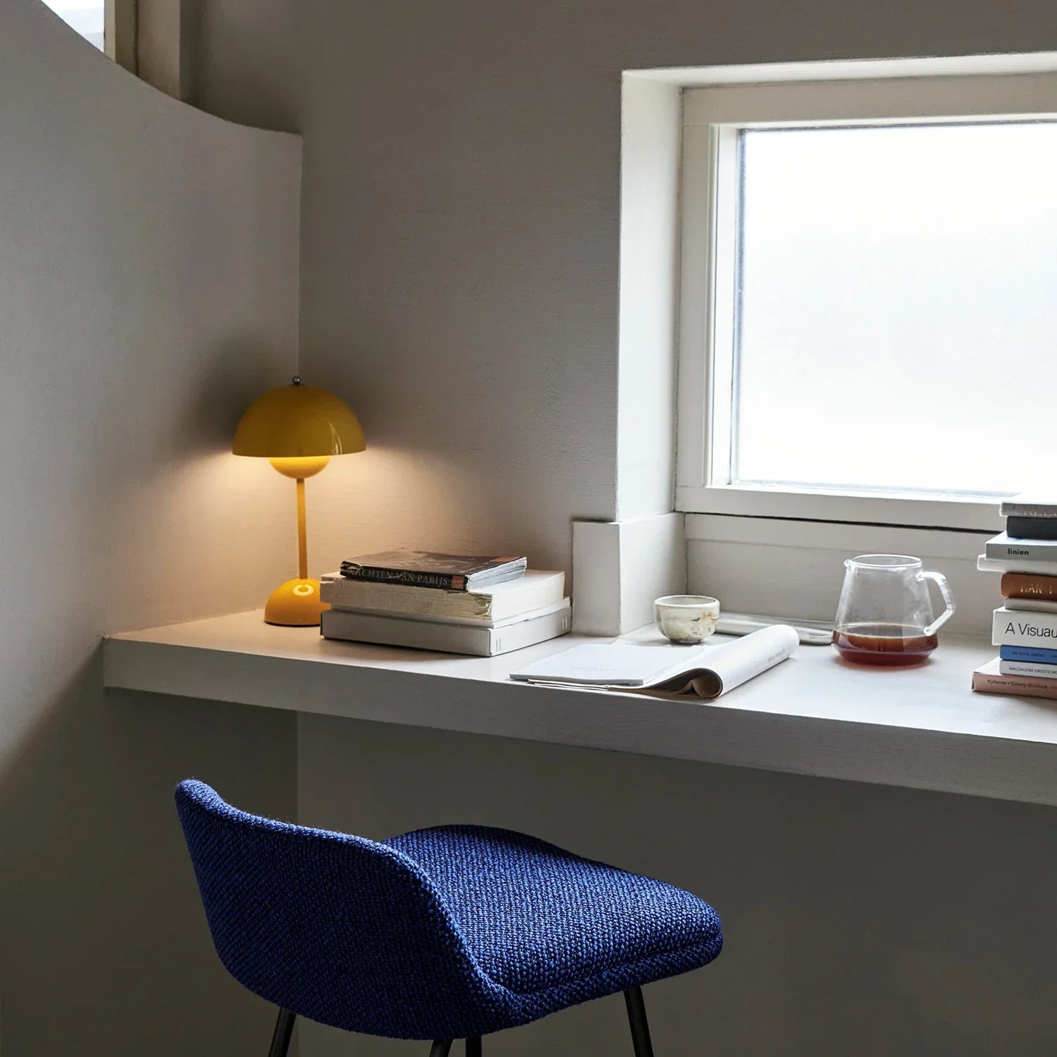 Yellow desk lamp on modern windowsill with books, coffee, and blue chair — cozy reading nook lighting